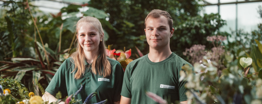 Zwei Personen stehen in einer grünen Umgebung, vermutlich einem Gewächshaus, umgeben von Pflanzen und Blumen.